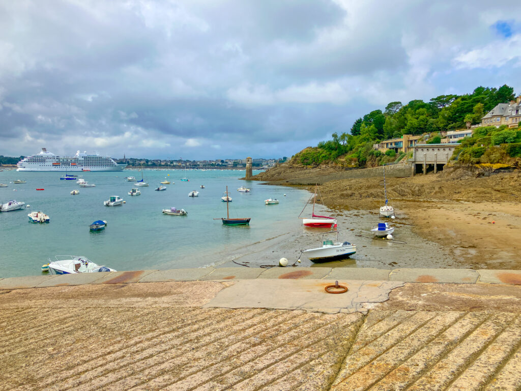 A coastal view featuring several small boats moored in a harbor, with a large cruise ship in the background and a rocky shoreline in the foreground, surrounded by lush greenery and residential buildings.