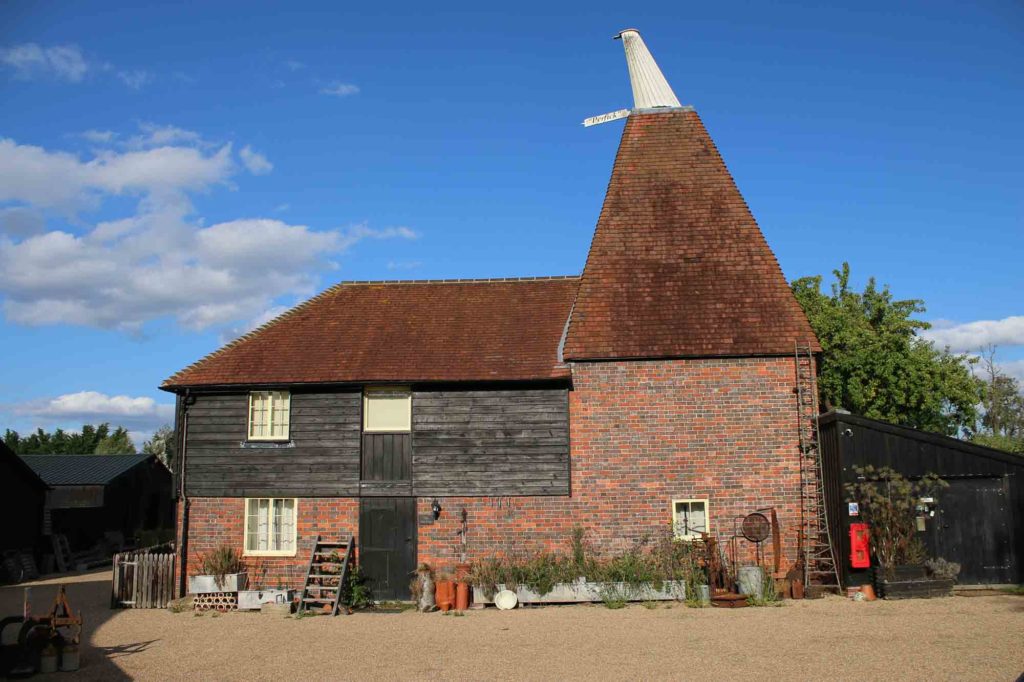 A traditional English oast house featuring a characteristic conical roof and a mix of brick and wooden exterior, set against a blue sky with scattered clouds.