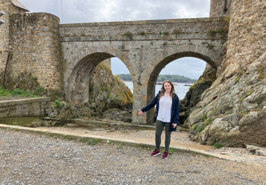 A woman stands beside a stone archway near a scenic harbor in Brittany, France, with rocky terrain and boats visible in the background.