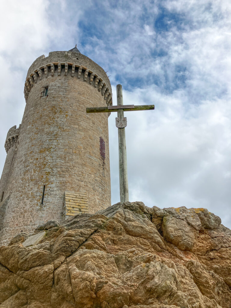 A stone tower with a cross standing on rocky terrain against a cloudy sky.